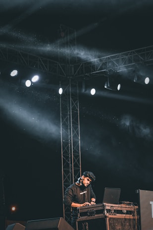 A person is performing on stage with a laptop and DJ equipment, under a framework of lights and cables. The scene is dark with bright spotlights creating beams of light and some haze filling the area. The person appears focused on their equipment.