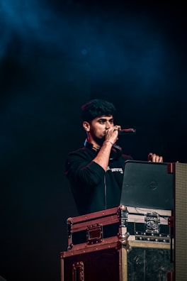 A person holding a microphone stands behind a DJ setup with a laptop and audio equipment. The background is dark with blue lighting, creating a concert or event atmosphere.