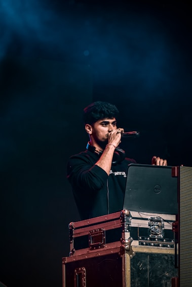 A person holding a microphone stands behind a DJ setup with a laptop and audio equipment. The background is dark with blue lighting, creating a concert or event atmosphere.