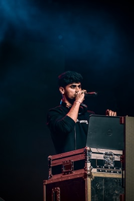 A person holding a microphone stands behind a DJ setup with a laptop and audio equipment. The background is dark with blue lighting, creating a concert or event atmosphere.