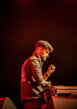 A person is standing behind a DJ mixing console, wearing headphones around their neck and a dark jacket with printed lettering on the back. Their right hand is raised as if adjusting sound levels. The background is dimly lit with a warm, reddish hue, creating a focused and intense atmosphere.