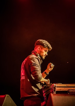 A person is standing behind a DJ mixing console, wearing headphones around their neck and a dark jacket with printed lettering on the back. Their right hand is raised as if adjusting sound levels. The background is dimly lit with a warm, reddish hue, creating a focused and intense atmosphere.