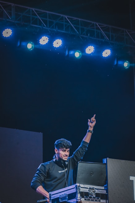 A DJ is performing on stage, looking energized and engaged with the music, as he raises one arm in the air. The scene is set under bright blue stage lights, and the DJ is standing behind a console with various equipment.