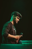 A person holding a microphone appears to be deep in thought or possibly preparing for a performance. The scene is dimly lit with a greenish hue surrounding the individual. The person is wearing a dark sweatshirt and is positioned beside a metal flight case.