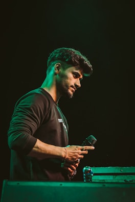 A person holding a microphone appears to be deep in thought or possibly preparing for a performance. The scene is dimly lit with a greenish hue surrounding the individual. The person is wearing a dark sweatshirt and is positioned beside a metal flight case.