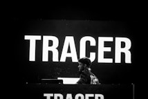 A person stands in front of a large electronic display with the word 'TRACER' illuminated in bold white letters. The background is predominantly dark, creating a high contrast with the bright text. The individual appears to be engaged with equipment, possibly related to music or a presentation.