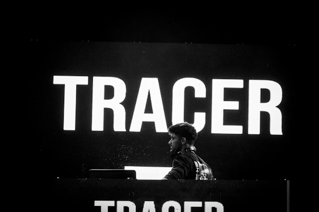 A person stands in front of a large electronic display with the word 'TRACER' illuminated in bold white letters. The background is predominantly dark, creating a high contrast with the bright text. The individual appears to be engaged with equipment, possibly related to music or a presentation.