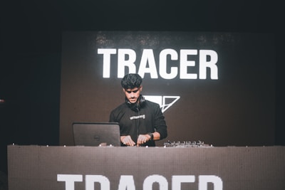 A person is performing as a DJ behind a large console with equipment. The background displays the word 'TRACER' in bold, white letters on a dark screen.