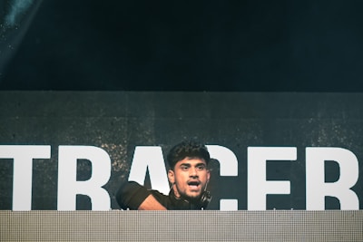 A person is on stage, positioned behind a large DJ booth or console. The background features large, illuminated letters, although the full text is not visible. The lighting is dim with a spotlight focused on the person, who appears to be engaged in performing or entertaining.