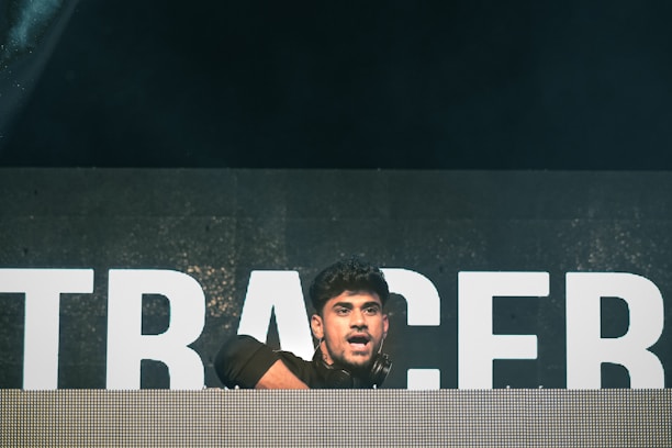 A person is on stage, positioned behind a large DJ booth or console. The background features large, illuminated letters, although the full text is not visible. The lighting is dim with a spotlight focused on the person, who appears to be engaged in performing or entertaining.