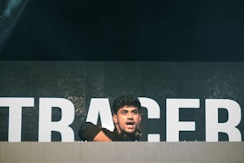 A person is on stage, positioned behind a large DJ booth or console. The background features large, illuminated letters, although the full text is not visible. The lighting is dim with a spotlight focused on the person, who appears to be engaged in performing or entertaining.