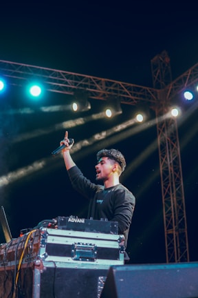 A DJ standing behind a setup with turntables and sound equipment, holding a microphone in one hand. Bright stage lights of varying colors illuminate the scene, with beams of light shining across the dark background. The person appears to be in an energetic stance.