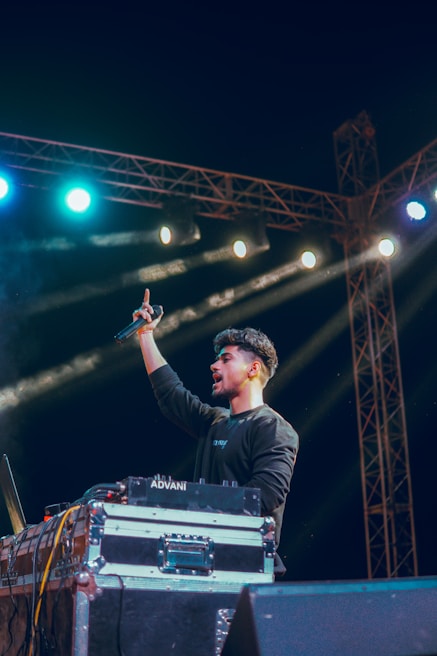 A DJ standing behind a setup with turntables and sound equipment, holding a microphone in one hand. Bright stage lights of varying colors illuminate the scene, with beams of light shining across the dark background. The person appears to be in an energetic stance.