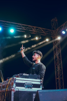 A DJ standing behind a setup with turntables and sound equipment, holding a microphone in one hand. Bright stage lights of varying colors illuminate the scene, with beams of light shining across the dark background. The person appears to be in an energetic stance.