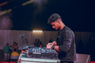 A person is operating DJ equipment on a stage with a laptop and a mixer, surrounded by dim ambient lighting. In the background, a group of people stand against a white curtain.