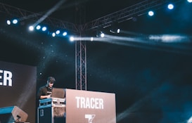 A DJ stands behind a console on a stage, surrounded by electronic equipment. Bright stage lights illuminate the scene, creating streaks of light against a dark background. A large screen behind the DJ displays the word 'TRACER'.