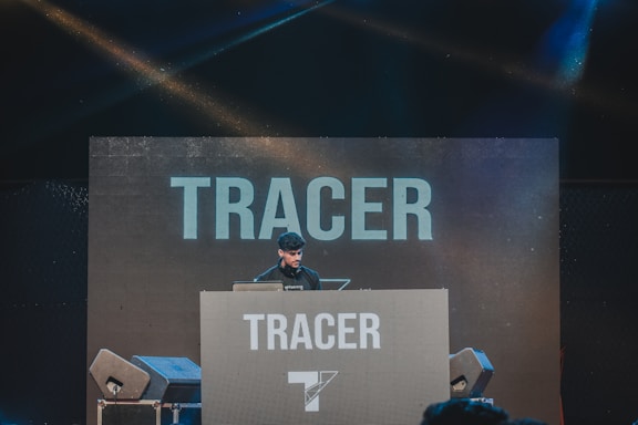 A DJ is performing on stage behind a large console with the word 'TRACER' displayed prominently on the backdrop and the console itself. Speakers are visible on either side, and colorful stage lights create a vibrant atmosphere.