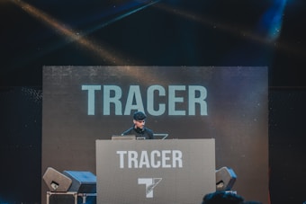 A DJ is performing on stage behind a large console with the word 'TRACER' displayed prominently on the backdrop and the console itself. Speakers are visible on either side, and colorful stage lights create a vibrant atmosphere.