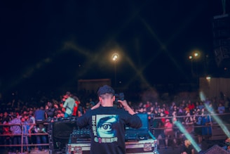 A person is standing at a DJ booth operating music equipment, with a large crowd gathered in front. The scene is illuminated with light beams creating an energetic nightclub or concert atmosphere. The person is wearing a shirt with a large eye graphic on the back, surrounded by text in a different language. The crowd appears engaged and lively.