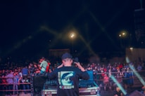 A person is standing at a DJ booth operating music equipment, with a large crowd gathered in front. The scene is illuminated with light beams creating an energetic nightclub or concert atmosphere. The person is wearing a shirt with a large eye graphic on the back, surrounded by text in a different language. The crowd appears engaged and lively.