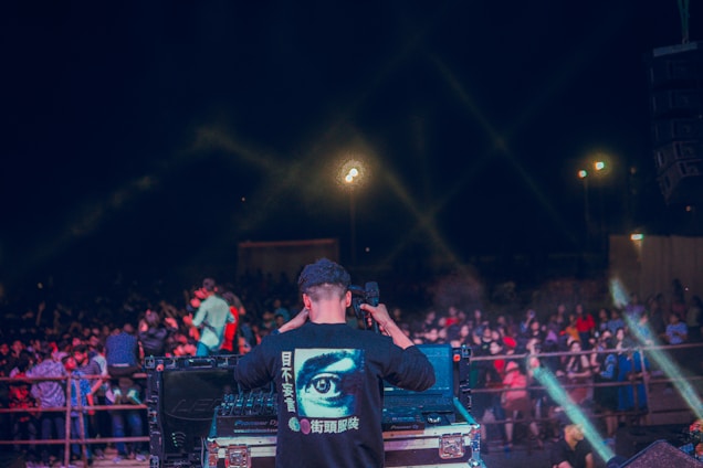 A person is standing at a DJ booth operating music equipment, with a large crowd gathered in front. The scene is illuminated with light beams creating an energetic nightclub or concert atmosphere. The person is wearing a shirt with a large eye graphic on the back, surrounded by text in a different language. The crowd appears engaged and lively.