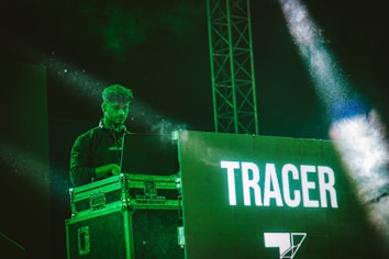 A person is standing behind a DJ booth, focused on a laptop while wearing headphones around their neck. The scene is illuminated with green lighting, creating a vibrant atmosphere. In the background, there is a large screen displaying the word 'TRACER'.