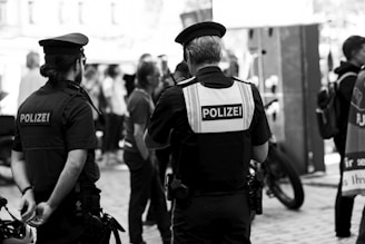 Two police officers are standing in an outdoor public area, facing away from the camera. They are wearing uniforms and have the word 'Polizei' visible on their vests. Several people are gathered around, some in casual attire, suggesting a crowd or public event. Bicycles are visible in the background.