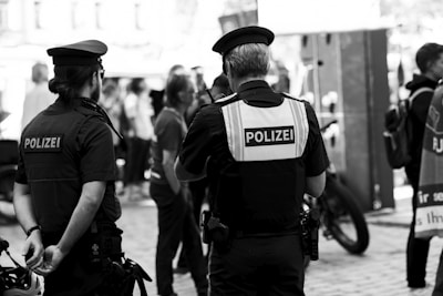 Two police officers are standing in an outdoor public area, facing away from the camera. They are wearing uniforms and have the word 'Polizei' visible on their vests. Several people are gathered around, some in casual attire, suggesting a crowd or public event. Bicycles are visible in the background.