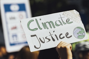 A cardboard sign with the words 'Climate Justice' written in green and black is being held up. The sign also features an illustration of the Earth. A date, '15.09.2023,' is visible on the top right corner of the sign. The background is blurred, with hints of other signs or people, suggesting a rally or protest.