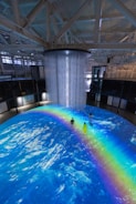 An indoor scene with a large circular projection on the floor depicting an ocean with a vibrant rainbow. Three people are standing on the projection, which creates an immersive effect. A cylindrical installation with lights hangs from the ceiling, adding to the atmosphere.