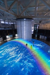 An indoor scene with a large circular projection on the floor depicting an ocean with a vibrant rainbow. Three people are standing on the projection, which creates an immersive effect. A cylindrical installation with lights hangs from the ceiling, adding to the atmosphere.