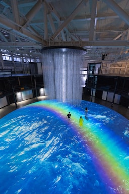 An indoor scene with a large circular projection on the floor depicting an ocean with a vibrant rainbow. Three people are standing on the projection, which creates an immersive effect. A cylindrical installation with lights hangs from the ceiling, adding to the atmosphere.