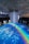 An indoor scene with a large circular projection on the floor depicting an ocean with a vibrant rainbow. Three people are standing on the projection, which creates an immersive effect. A cylindrical installation with lights hangs from the ceiling, adding to the atmosphere.