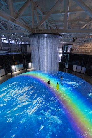 An indoor scene with a large circular projection on the floor depicting an ocean with a vibrant rainbow. Three people are standing on the projection, which creates an immersive effect. A cylindrical installation with lights hangs from the ceiling, adding to the atmosphere.