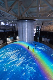 An indoor scene with a large circular projection on the floor depicting an ocean with a vibrant rainbow. Three people are standing on the projection, which creates an immersive effect. A cylindrical installation with lights hangs from the ceiling, adding to the atmosphere.