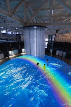 An indoor scene with a large circular projection on the floor depicting an ocean with a vibrant rainbow. Three people are standing on the projection, which creates an immersive effect. A cylindrical installation with lights hangs from the ceiling, adding to the atmosphere.