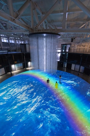 An indoor scene with a large circular projection on the floor depicting an ocean with a vibrant rainbow. Three people are standing on the projection, which creates an immersive effect. A cylindrical installation with lights hangs from the ceiling, adding to the atmosphere.