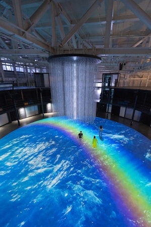 An indoor scene with a large circular projection on the floor depicting an ocean with a vibrant rainbow. Three people are standing on the projection, which creates an immersive effect. A cylindrical installation with lights hangs from the ceiling, adding to the atmosphere.