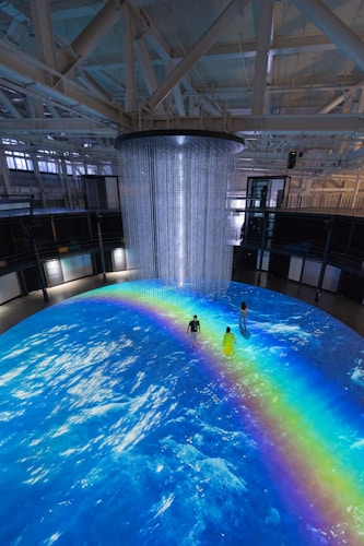 An indoor scene with a large circular projection on the floor depicting an ocean with a vibrant rainbow. Three people are standing on the projection, which creates an immersive effect. A cylindrical installation with lights hangs from the ceiling, adding to the atmosphere.