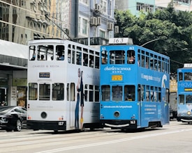 Two double-decker trams, one white and one blue, travel alongside each other on a city street. The trams display advertisements for different brands. There are several passengers visible inside the trams, and buildings are seen in the background.