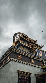 An ornate and traditional structure with intricate carvings and decorations, featuring a large roof adorned with gold and colorful details. The building includes architectural elements typical of monastic or religious significance, with a prominent image of a person displayed on one side. A cloudy sky serves as the backdrop, adding a somber tone to the scene.