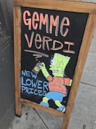A wooden-framed chalkboard display on a sidewalk featuring colorful hand-drawn artwork. The text 'GEMME VERDI' is written in large, bold letters at the top in an orange hue. Below, 'NEW LOWER PRICES' is depicted in blue, next to a cartoon character wearing an orange shirt and blue shorts, holding a can.