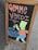 A wooden-framed chalkboard display on a sidewalk featuring colorful hand-drawn artwork. The text 'GEMME VERDI' is written in large, bold letters at the top in an orange hue. Below, 'NEW LOWER PRICES' is depicted in blue, next to a cartoon character wearing an orange shirt and blue shorts, holding a can.
