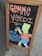A wooden-framed chalkboard display on a sidewalk featuring colorful hand-drawn artwork. The text 'GEMME VERDI' is written in large, bold letters at the top in an orange hue. Below, 'NEW LOWER PRICES' is depicted in blue, next to a cartoon character wearing an orange shirt and blue shorts, holding a can.