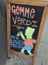 A wooden-framed chalkboard display on a sidewalk featuring colorful hand-drawn artwork. The text 'GEMME VERDI' is written in large, bold letters at the top in an orange hue. Below, 'NEW LOWER PRICES' is depicted in blue, next to a cartoon character wearing an orange shirt and blue shorts, holding a can.