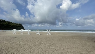 Large letters spelling out 'RELAX' are positioned on a sandy beach with waves in the background. The sky is partly cloudy, and there is lush greenery on the left side.