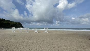 Large letters spelling out 'RELAX' are positioned on a sandy beach with waves in the background. The sky is partly cloudy, and there is lush greenery on the left side.