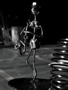 A metallic sculpture of a musician holding a saxophone, crafted from nuts and bolts, stands prominently against a dark background. The figure is sleek and abstract, with elongated limbs and a small, rounded head. Nearby, a shiny metal coil adds an industrial feel. The scene is dimly lit, creating striking reflections on the metal surfaces.