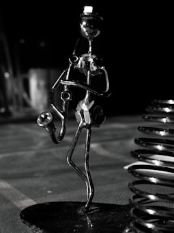 A metallic sculpture of a musician holding a saxophone, crafted from nuts and bolts, stands prominently against a dark background. The figure is sleek and abstract, with elongated limbs and a small, rounded head. Nearby, a shiny metal coil adds an industrial feel. The scene is dimly lit, creating striking reflections on the metal surfaces.
