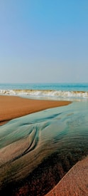 A serene beach scene with gentle waves rolling onto the sandy shore. Clear blue sky stretches across the horizon. A small stream of water creates patterns in the wet sand as it makes its way back to the ocean.